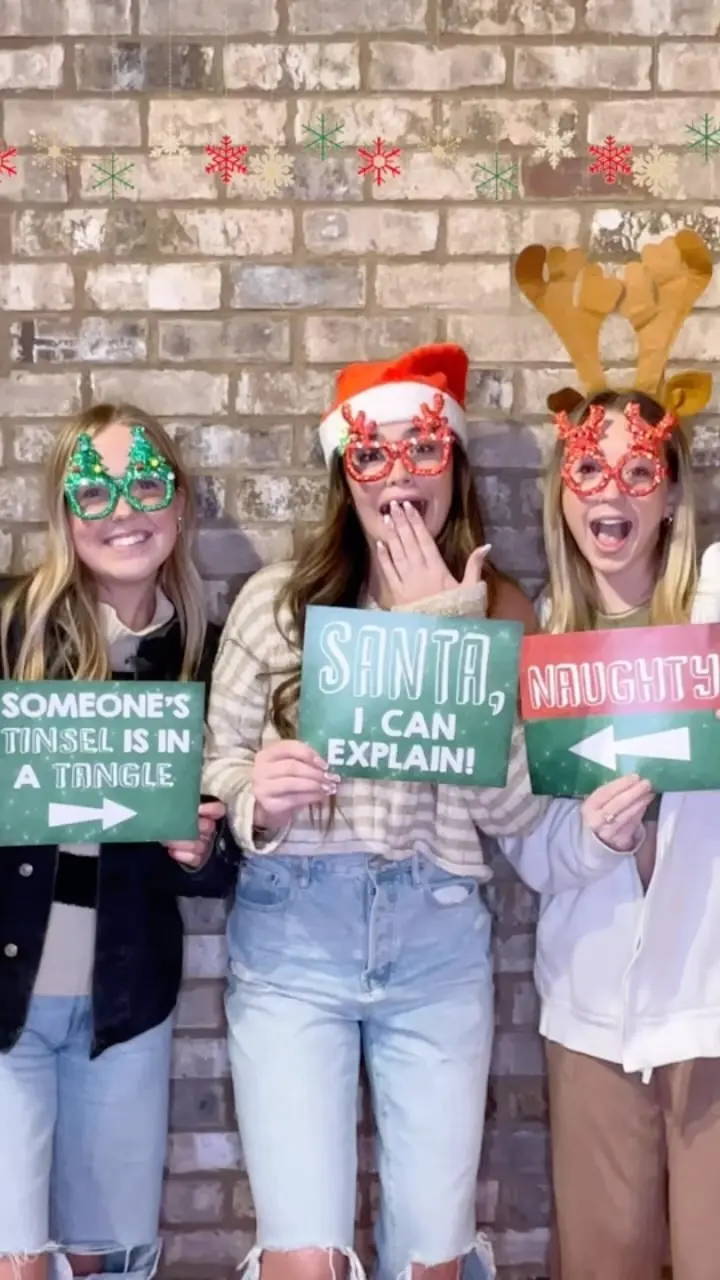 Three guests with festive glasses holding holiday signs at a brick-wall photo booth
