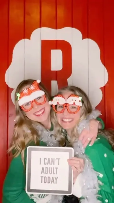 Two friends in Santa glasses holding an 'I Can't Adult Today' sign